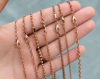 Op de afbeelding: Een verzameling van zes roségoudkleurige metalen kettingen in verschillende stijlen, waaronder bolletjes-, box- en schakelontwerpen. Elke ketting is voorzien van een karabijnsluiting en toont diverse texturen en patronen in metaal.