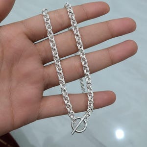 Puede incluir: Un collar de cadena de tono plateado que presenta una serie de eslabones circulares entrelazados que crean una apariencia densa y texturizada. El collar se asegura con un cierre de palanca que consiste en un anillo y una barra metálica.