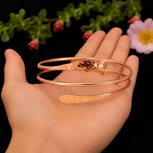 Op de afbeelding: Een koperkleurige metalen armband met een spiraalvormig ontwerp en twee overlappende uiteinden. De uiteinden van de armband zijn afgeplat en hebben een getextureerde, gehamerde afwerking, terwijl de rest van de band glad en rond is.