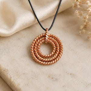 Könnte beinhalten: Ein roségoldfarbener Anhänger bestehend aus drei konzentrischen Kreisen aus gedrehtem, seilartigem Metall. Der Anhänger hängt an einer dünnen schwarzen Kordel. Das Design besteht aus drei ineinander verschlungenen Ringen unterschiedlicher Größe.