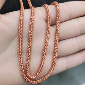 Op de afbeelding: Een koperkleurige tarweketting met een strak geweven, gevlochten metalen schakelontwerp. De ketting heeft een duidelijke metaalglans en een afgeronde, flexibele structuur die over zichzelf heen lussen vormt en het patroon toont.