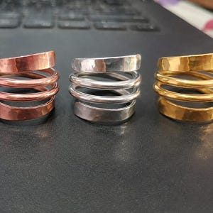Puede incluir: Tres anillos en espiral dispuestos en fila, con un anillo de tono cobre a la izquierda, un anillo de tono plateado en el centro y un anillo de tono dorado a la derecha. Cada anillo tiene una textura martillada en la banda plana y un diseño de múltiples vueltas.