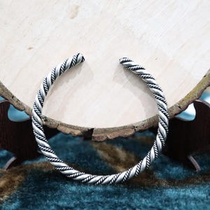 May include: A silver-toned metal cuff bracelet featuring a continuous twisted rope texture. The bracelet is an open-ended circular design with a polished finish that highlights the intricate spiral pattern along the entire length of the band.