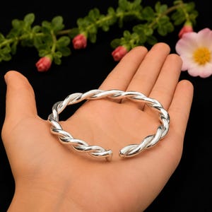 Può includere: Un bracciale a fascia in argento lucido caratterizzato da un design a corda intrecciata. Il bracciale è un anello circolare aperto con una finitura metallica liscia e riflettente, che presenta un motivo a spirale continuo lungo tutto il gioiello.