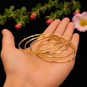 Può includere: Un set di cinque braccialetti circolari sottili in oro, caratterizzati da una texture martellata e sfaccettata che riflette la luce. I braccialetti sono impilati, mostrando una finitura giallo metallizzato brillante e un design sottile.
