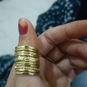 Può includere: Una pila di sette anelli in metallo color oro indossati su un pollice. Gli anelli presentano vari design, tra cui fasce lisce lucide, una fascia con un motivo floreale o a foglie ripetuto e una fascia con dettagli testurizzati o dentellati.