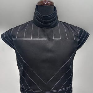 Peut inclure: Un haut noir sans manches doté d'un col montant avec des surpiqûres horizontales. Le vêtement est orné de fines rayures blanches formant des lignes verticales sur le haut de la poitrine et des motifs angulaires en chevron sur le bas.