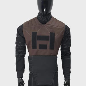 May include: A brown and black tactical vest featuring horizontal quilted padding, three rectangular black hook-and-loop fastener patches on the chest, and a high collar, worn over a black long-sleeved shirt with white decorative stitching details.