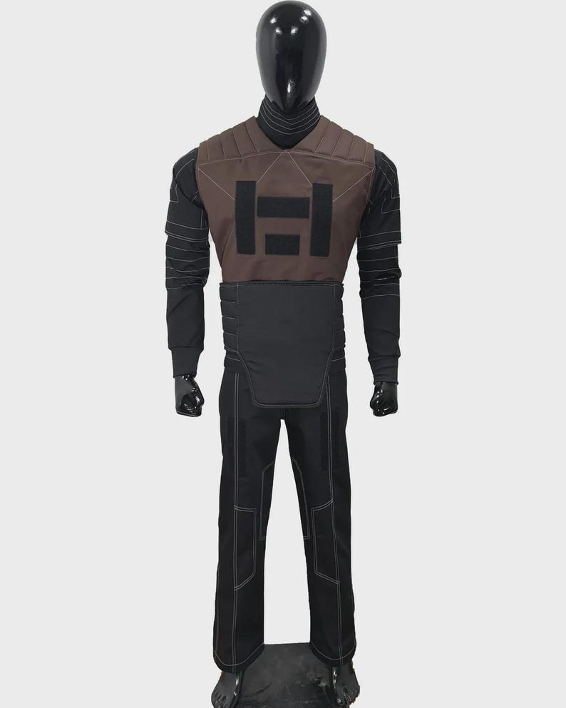 May include: A full-body costume featuring a black long-sleeved shirt with white stitching, a brown chest piece with a black rectangular graphic, and a black waist guard. The outfit includes matching black trousers with white seam detailing.