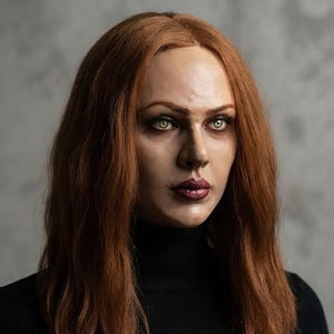May include: A realistic mannequin head featuring long, wavy auburn hair, light green eyes, and dark plum-colored lips. The mannequin has a smooth, skin-toned complexion and is wearing a black high-neck garment that covers the neck and shoulders.
