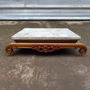 May include: A rectangular display stand featuring a polished light brown wooden base with curved scroll feet and intricate carved detailing at the center. The top surface consists of a flat, light gray stone slab with natural veining patterns.