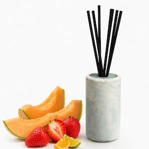 Peut inclure: Un diffuseur cylindrique en marbre bleu clair et blanc contenant cinq bâtonnets noirs fins, placé à côté de deux tranches de melon cantaloup orange, deux fraises entières, une fraise coupée en deux et deux petits quartiers de citron vert.