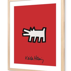 Pode incluir: Uma impressão de arte emoldurada apresentando uma ilustração de um cão branco estilizado com contornos pretos grossos centralizada num fundo vermelho sólido. A assinatura Keith Haring está escrita em letra preta na parte inferior do campo vermelho dentro de uma moldura de madeira clara.