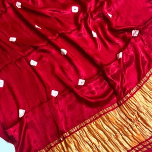May include: A vibrant red fabric featuring a scattered pattern of small white square-shaped tie-dye accents. The bottom edge is finished with a wide, textured gold metallic border that has a crinkled appearance and horizontal striped detailing.