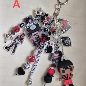 May include: A decorative keychain featuring a silver ring and multiple dangling charms, including a small vinyl record, various acrylic portraits of a performer, a miniature figurine in a red outfit, and beads in black, white, and red with the text KING OF POP.