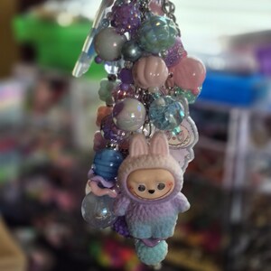 Op de afbeelding: Een decoratieve sleutelhanger met een verscheidenheid aan pastelkleurige kralen in tinten paars, roze en blauw. De hanger bevat een klein, pluizig figuurtje met een capuchon met konijnenoren en een klein hartvormig hangertje.
