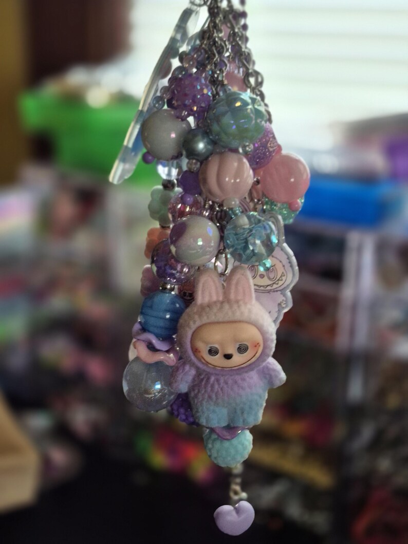 Op de afbeelding: Een decoratieve sleutelhanger met een verscheidenheid aan pastelkleurige kralen in tinten paars, roze en blauw. De hanger bevat een klein, pluizig figuurtje met een capuchon met konijnenoren en een klein hartvormig hangertje.