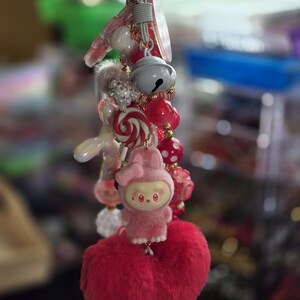 Op de afbeelding: Een decoratieve sleutelhanger met diverse bedels, waaronder een pluizig roze hart, een klein roze konijntje, een wit belletje, een rood-witte spiraalvormige snoepbedel en diverse rode, witte en roze kralen aan een metalen ringbevestiging.