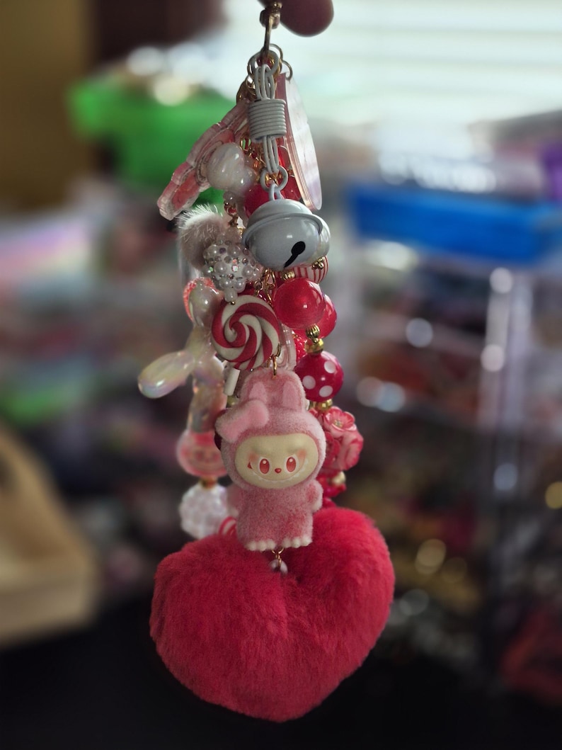 Op de afbeelding: Een decoratieve sleutelhanger met diverse bedels, waaronder een pluizig roze hart, een klein roze konijntje, een wit belletje, een rood-witte spiraalvormige snoepbedel en diverse rode, witte en roze kralen aan een metalen ringbevestiging.