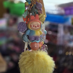 Op de afbeelding: Een decoratieve sleutelhanger met een centrale figuur van een glimlachend wezen met konijnenoren, gekleed in een veelkleurig pluizig pak. De hanger is versierd met diverse kralen, een paarse koekvormige bedel en een grote, pluizige gele pompon.