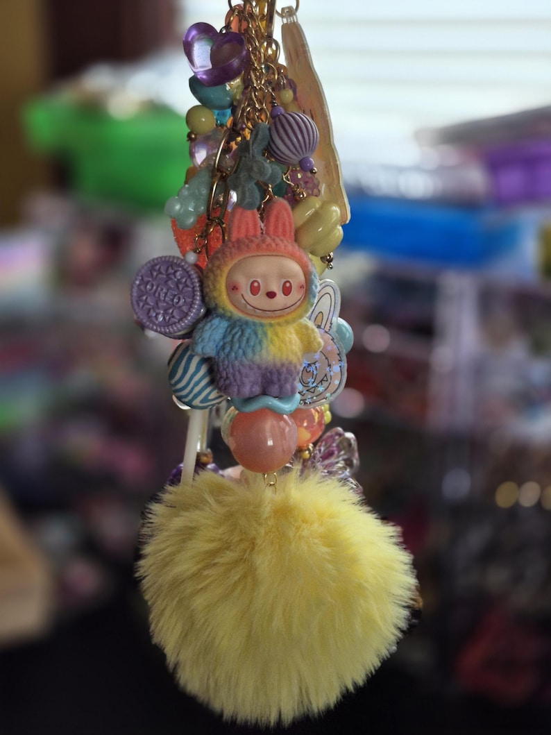 Op de afbeelding: Een decoratieve sleutelhanger met een centrale figuur van een glimlachend wezen met konijnenoren, gekleed in een veelkleurig pluizig pak. De hanger is versierd met diverse kralen, een paarse koekvormige bedel en een grote, pluizige gele pompon.