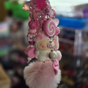 Op de afbeelding: Een decoratieve sleutelhanger met diverse roze kralen, waaronder hartvormen, een spiraalvormige lollybedel en een klein wit figuurtje. Het accessoire is afgewerkt met een zilveren belletje en een zachte, pluizige lichtroze pompon aan de onderzijde.