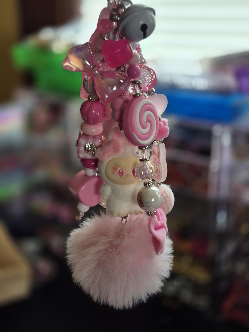 Op de afbeelding: Een decoratieve sleutelhanger met diverse roze kralen, waaronder hartvormen, een spiraalvormige lollybedel en een klein wit figuurtje. Het accessoire is afgewerkt met een zilveren belletje en een zachte, pluizige lichtroze pompon aan de onderzijde.