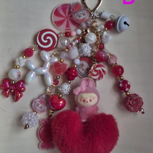 Op de afbeelding: Een decoratieve sleutelhanger met een roze pluizig hart, een klein roze konijnenfiguurtje en diverse bedels, waaronder rood-witte spiraalsnoepjes, iriserende ballonvormige kralen, rode strikken en een wit belletje aan een hartvormige metalen ring.