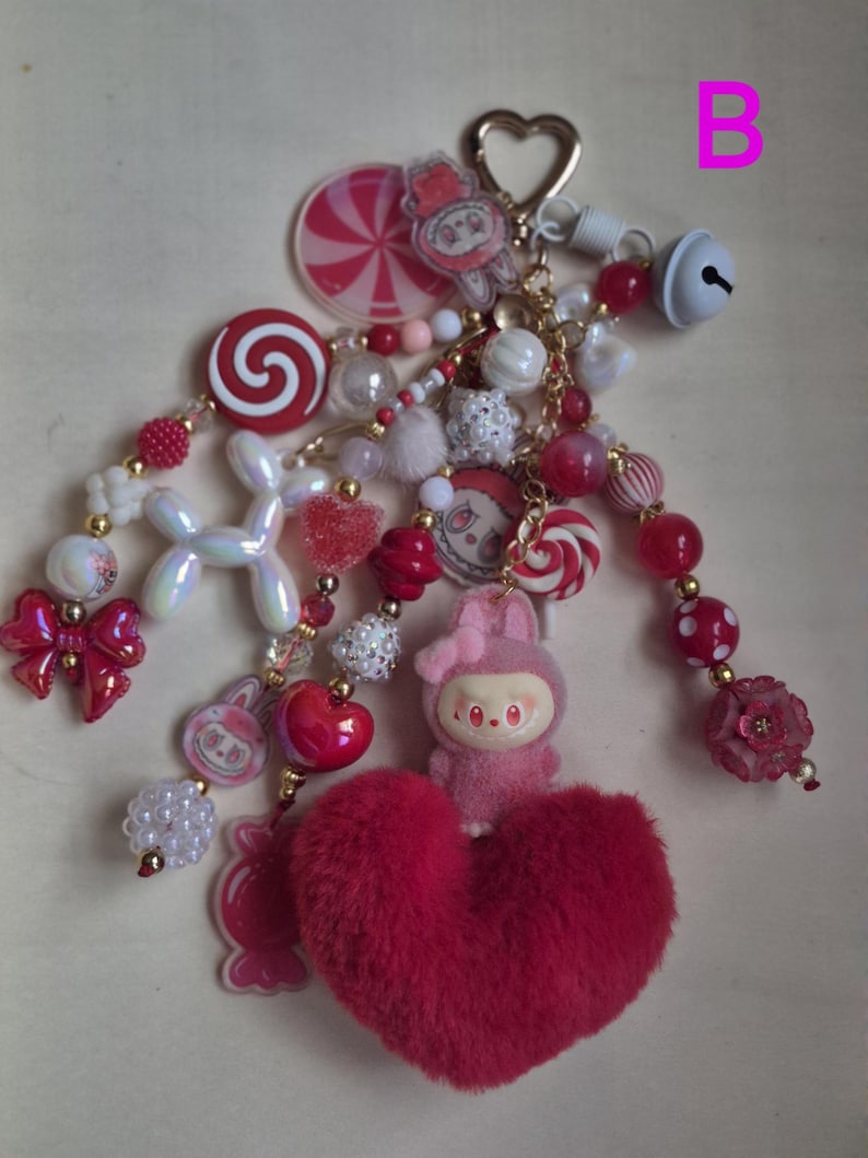 Op de afbeelding: Een decoratieve sleutelhanger met een roze pluizig hart, een klein roze konijnenfiguurtje en diverse bedels, waaronder rood-witte spiraalsnoepjes, iriserende ballonvormige kralen, rode strikken en een wit belletje aan een hartvormige metalen ring.