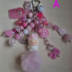 Op de afbeelding: Een decoratieve sleutelhanger met een hartvormige metalen sluiting, een wit belletje en diverse roze en witte kralen. Het accessoire bevat bedels in de vorm van een konijn, een spiraallolly en een strik, afgewerkt met een zachte roze pompon.