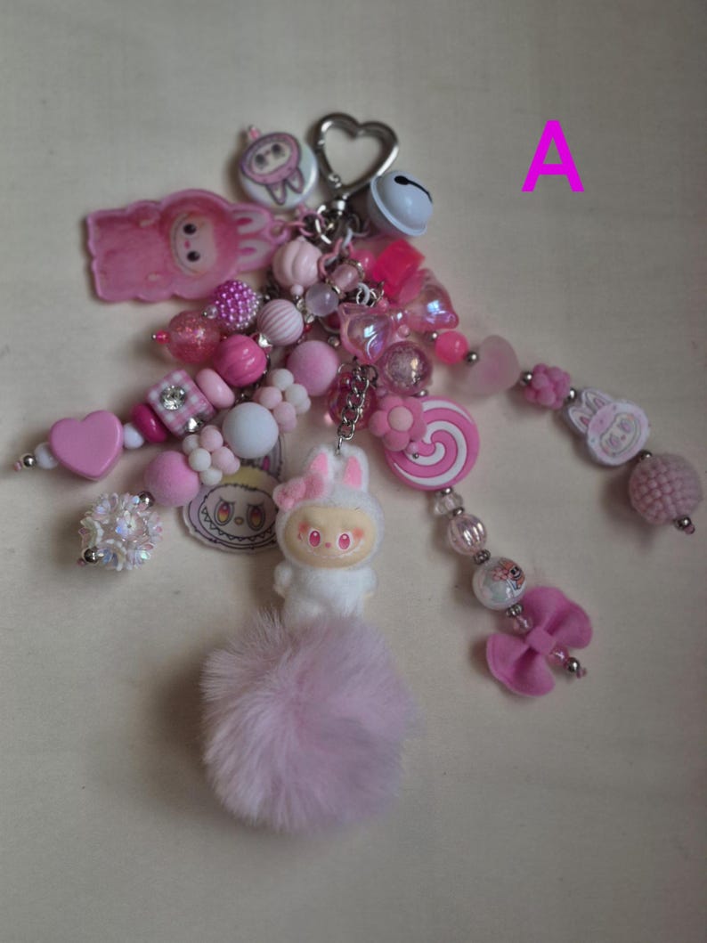 Op de afbeelding: Een decoratieve sleutelhanger met een hartvormige metalen sluiting, een wit belletje en diverse roze en witte kralen. Het accessoire bevat bedels in de vorm van een konijn, een spiraallolly en een strik, afgewerkt met een zachte roze pompon.