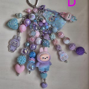 Op de afbeelding: Een kralen sleutelhanger met meerdere strengen, een zilveren hartvormige sluiting, diverse paarse, roze en blauwe kralen, en verschillende bedels waaronder een pluizig konijnenfiguurtje, een glinsterende blauwe wolk en platte acryl konijnhangers.