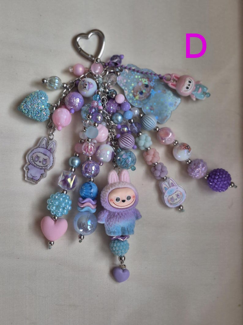 Op de afbeelding: Een kralen sleutelhanger met meerdere strengen, een zilveren hartvormige sluiting, diverse paarse, roze en blauwe kralen, en verschillende bedels waaronder een pluizig konijnenfiguurtje, een glinsterende blauwe wolk en platte acryl konijnhangers.