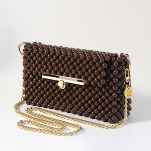 May include: A rectangular handbag constructed from dark brown spherical beads, featuring a gold-tone metal bar clasp closure and a matching gold-tone chain shoulder strap. A small circular gold tag with a bag icon and the text Merit Bags is attached.