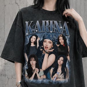 Puede incluir: Una camiseta gris carbón de corte oversize con un estampado gráfico de estilo vintage y el texto KARINA en letras azules. El diseño incluye varios retratos fotográficos de una persona joven en diversas poses sobre un fondo azul nublado.