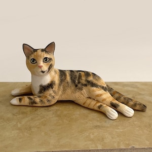 May include: A calico cat figurine in a reclining position, featuring a coat with patches of orange, black, and white. The cat has alert ears, dark eyes, and a white chest, with its front paws extended forward and its tail resting behind it.