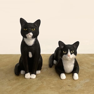 May include: Two black and white cat figurines with yellow eyes. One cat is in a sitting position, while the other cat is in a lying down position. Both figurines feature white chest patches, white paws, and detailed black fur texture throughout.