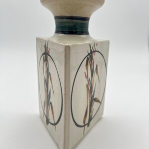 May include: A square, off-white ceramic vase with a flared top. The vase features a botanical design within black circular frames. The design includes brown, green, and pink floral elements. A dark green and black band encircles the neck.