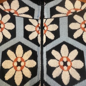 May include: Close-up of a zip-up sweater with a floral pattern. The sweater features a black background with light blue and black geometric shapes. Cream-colored flowers with orange centers are evenly spaced across the fabric. The zipper is black.