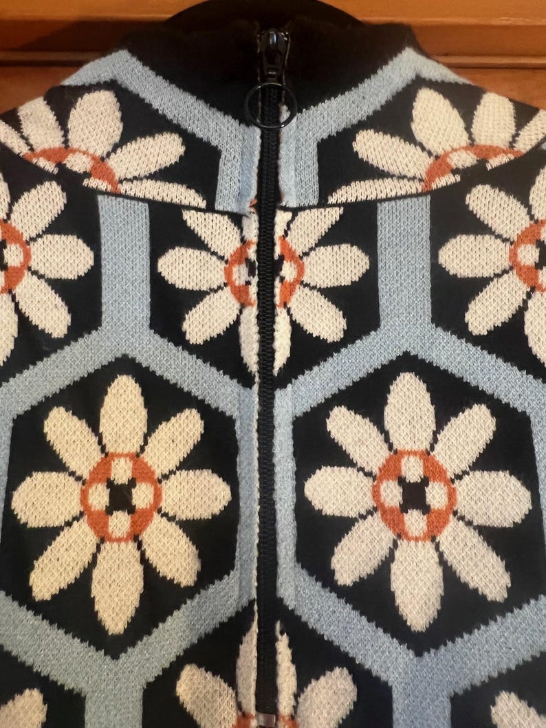 May include: Close-up of a zip-up sweater with a floral pattern. The sweater features a black background with light blue and black geometric shapes. Cream-colored flowers with orange centers are evenly spaced across the fabric. The zipper is black.