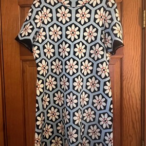 May include: A short-sleeved dress with a floral pattern. The dress features a high neckline and a geometric design of white flowers with red centers set against a light blue background. Black trim outlines the sleeves and hem.