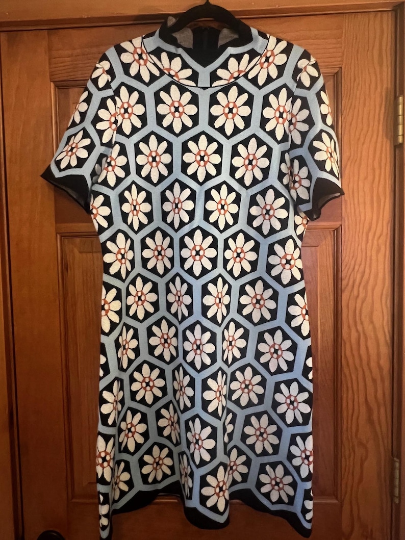 May include: A short-sleeved dress with a floral pattern. The dress features a high neckline and a geometric design of white flowers with red centers set against a light blue background. Black trim outlines the sleeves and hem.