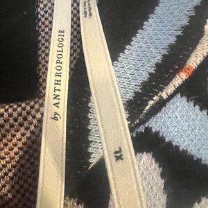 May include: Close-up of a sweater with a black, blue, and white pattern. The tag reads "by ANTHROPOLOGIE" and "XL". The sweater has a textured knit design. The tag also says "made in china".