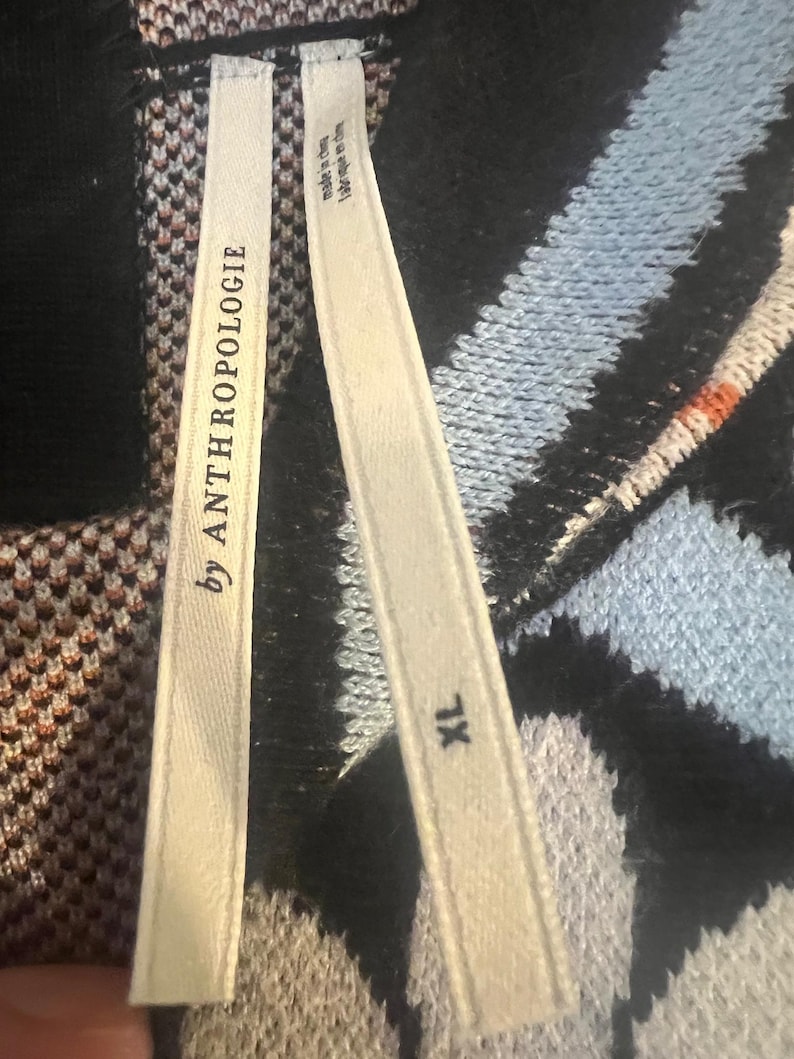 May include: Close-up of a sweater with a black, blue, and white pattern. The tag reads "by ANTHROPOLOGIE" and "XL". The sweater has a textured knit design. The tag also says "made in china".