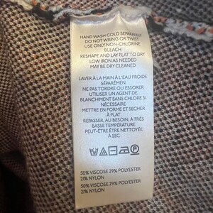 May include: A close-up of a clothing tag with care instructions and fabric composition details. The tag is white with black text, and the background is a brown and orange patterned fabric. The tag includes washing, drying, and ironing instructions.