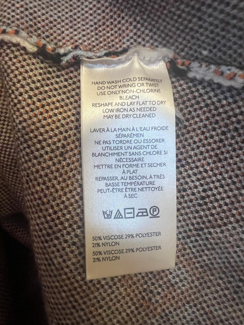 May include: A close-up of a clothing tag with care instructions and fabric composition details. The tag is white with black text, and the background is a brown and orange patterned fabric. The tag includes washing, drying, and ironing instructions.