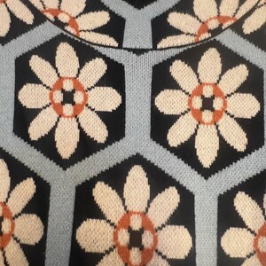 May include: Close-up of a knitted sweater featuring a repeating floral pattern. The design includes large white flowers with orange and black centers set against a black hexagonal background, outlined by light blue lines.
