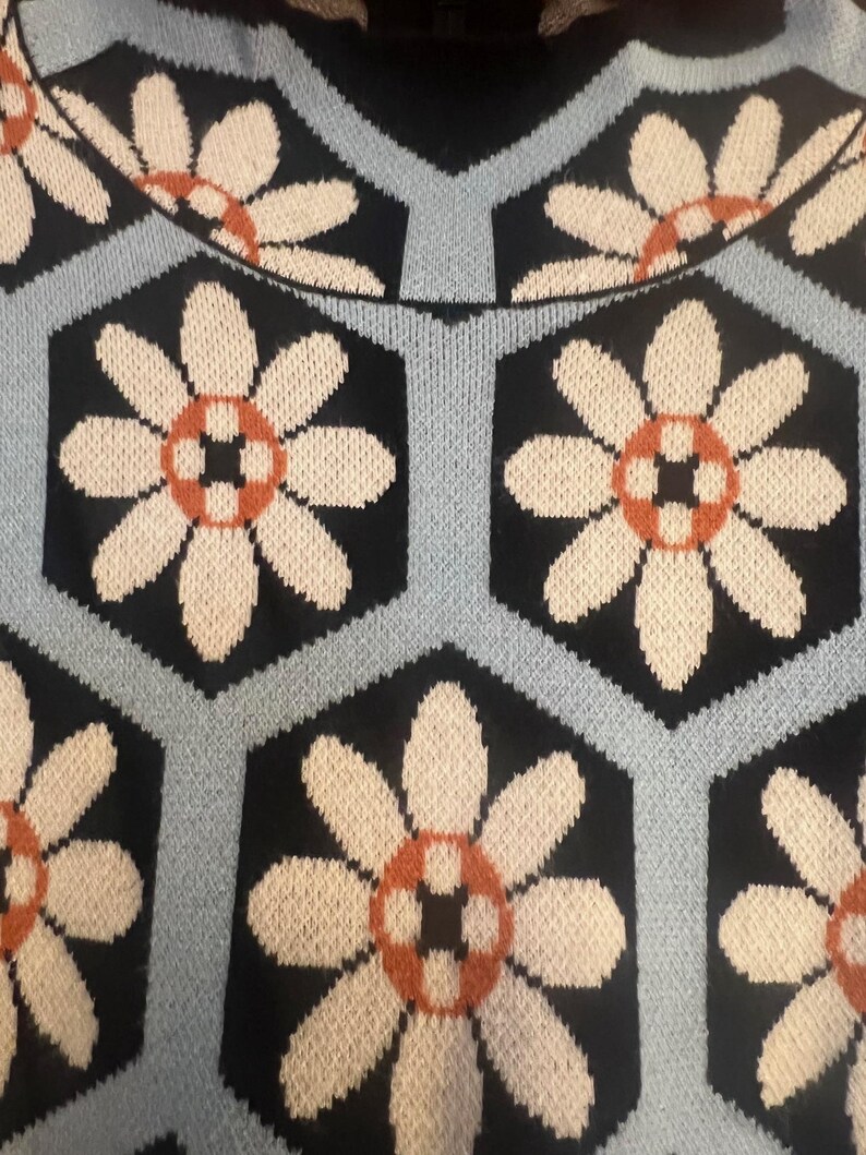 May include: Close-up of a knitted sweater featuring a repeating floral pattern. The design includes large white flowers with orange and black centers set against a black hexagonal background, outlined by light blue lines.