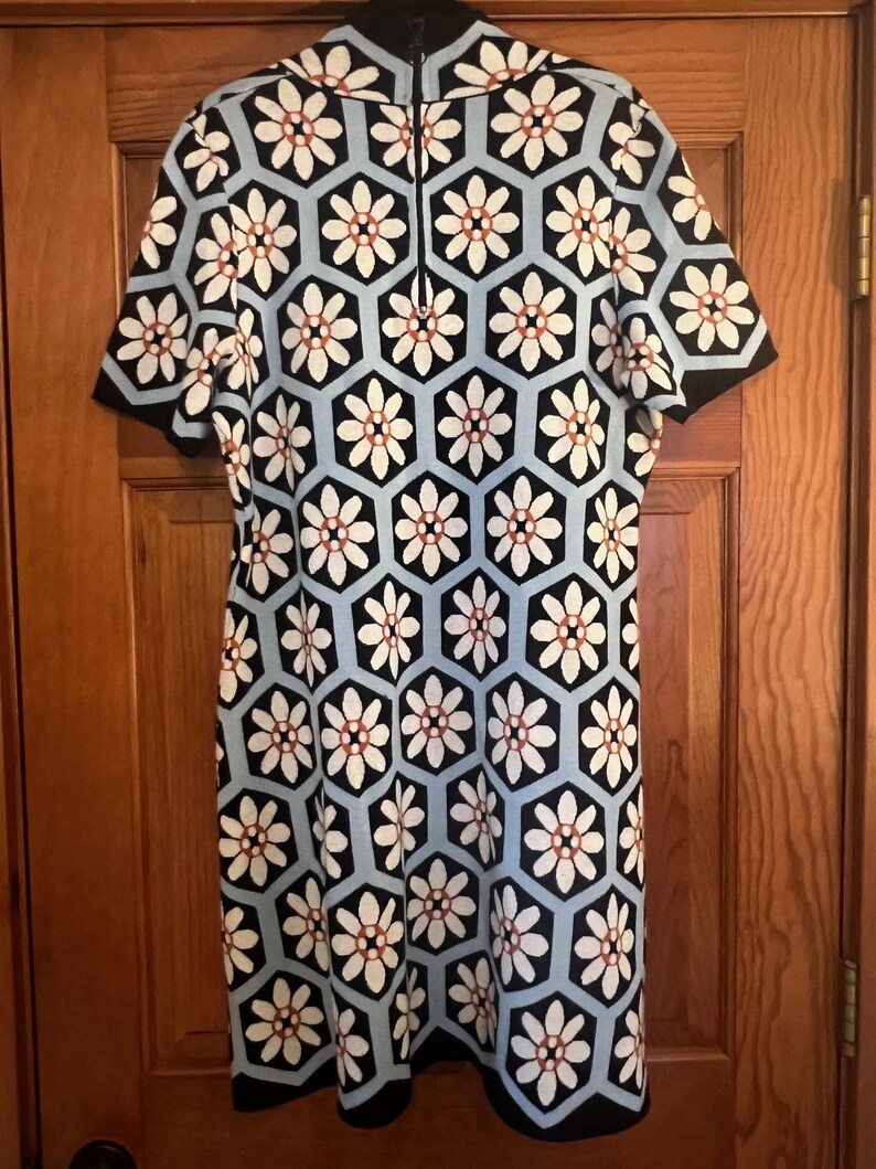 May include: A short-sleeved dress featuring a geometric floral pattern. The dress has a black, white, and light blue color scheme. The flowers are white with a brown center, set within black and light blue hexagons.