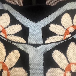 May include: Close-up of a black sweater with a floral pattern. The design features large white and orange flowers with light blue accents. The sweater has a zipper at the neckline and a textured knit.